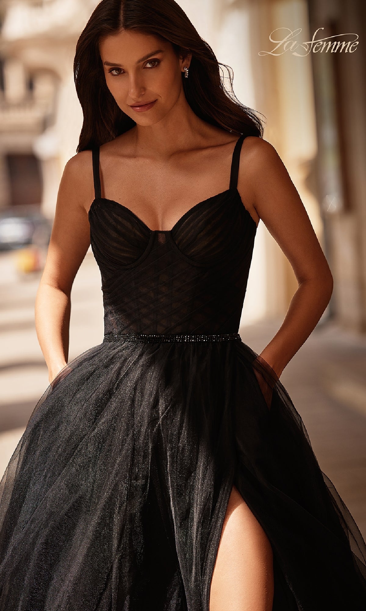 Woman in a sleeveless black La Femme 32956 beaded-waist prom ball gown with corset bodice and tulle skirt, standing outdoors with hands in pockets.